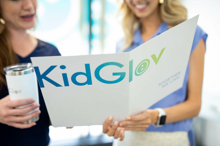 Two women look at a folder with the KidGlov logo featured prominently.