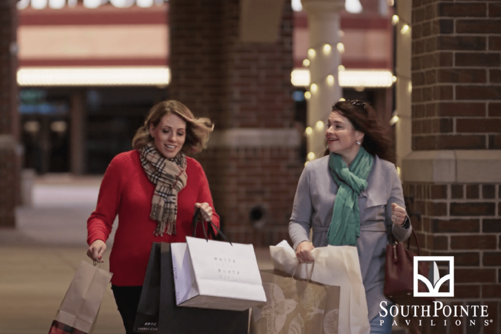 Two women walk through SouthPointe Pavilions at Christmas time, their arms are full of bags.