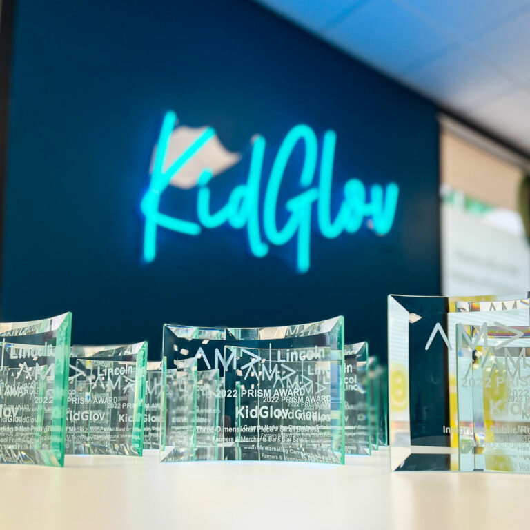 A close up shot of a row of glass AMA 2022 Prism Awards lined up on a desk in front of the KidGlov neon sign.