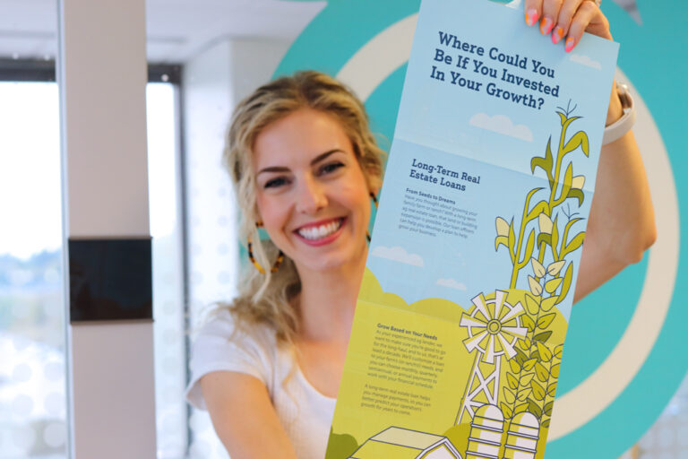 A blonde KidGlov employee smiles as she shows off a printed mailer for Farmers and Merchants Bank.