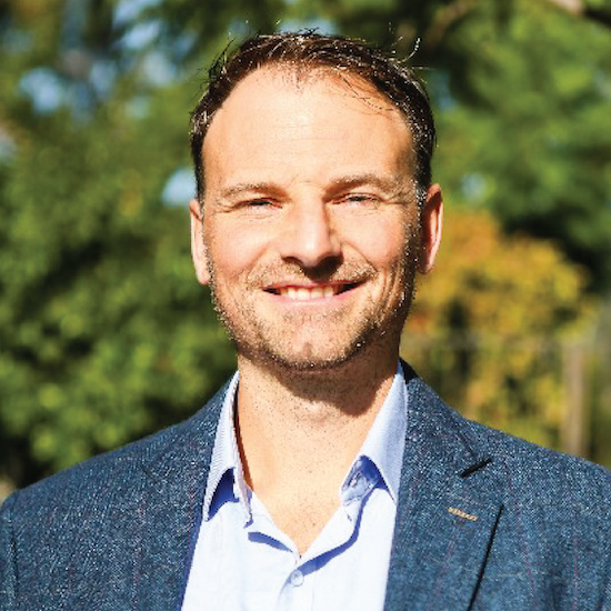 Headshot of Gareth Evans wearing a blue suit jacket over a button-down shirt while standing in front trees.