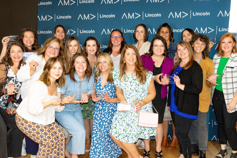 A group of women gather together while holding AMA Prism Awards. They stand in front of a blue step and repeat background that reads "AMA Lincoln".