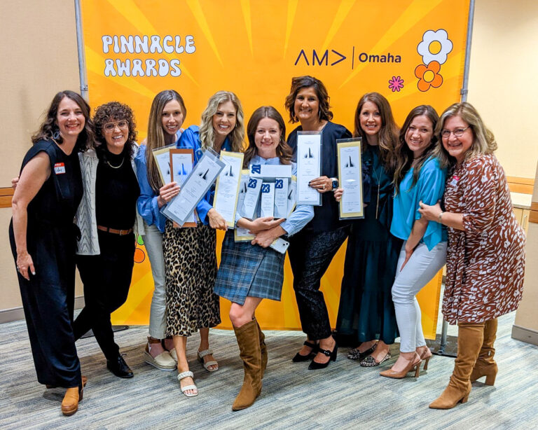 A group of women gather together smiling while holding AMA Pinnacle Awards. They stand in front of a yellow backdrop that reads "Pinnacle Awards, AMA Omaha".